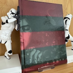Puede incluir: Un libro de tapa dura con una cubierta a rayas rojas y verdes, que presenta un primer plano de una cara y manos. El libro está sostenido por un soporte de plástico transparente, flanqueado por dos figuras blancas de Stormtroopers.