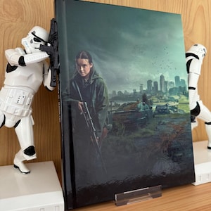 Puede incluir: Un libro de tapa dura con una cubierta brillante que muestra a una mujer con un rifle, ambientada en un paisaje urbano postapocalíptico. El libro está sostenido por dos figuras blancas de Stormtroopers, que actúan como sujetalibros. El libro está sobre una superficie de madera.