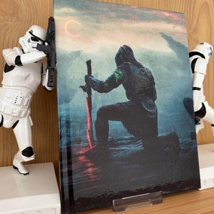 Puede incluir: Un libro de arte de fantasía con una figura arrodillada sosteniendo una espada roja brillante. La portada tiene una paleta de colores oscura y atmosférica con un cuerpo celeste en el cielo. Dos figuras blancas de Stormtroopers flanquean el libro.