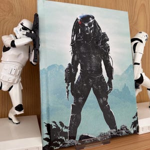 Puede incluir: Un libro de tapa dura con una ilustración del personaje Predator, sobre un fondo azul y verde. El libro está sostenido por dos figuras blancas de Stormtroopers sobre bases blancas.