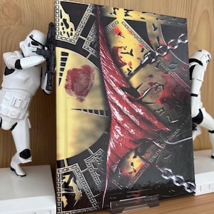 Puede incluir: Un libro de tapa dura con un diseño de portada llamativo, con una huella dactilar en forma de corazón y un elemento de tela roja rasgada. El libro está flanqueado por dos figuras blancas de Stormtroopers.