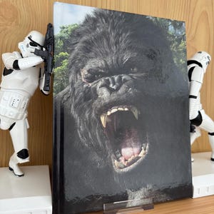 Puede incluir: Un libro de tapa dura con una imagen de primer plano de un gorila rugiendo. El libro está sostenido por dos sujetalibros blancos con forma de Stormtroopers, sobre una superficie de madera.