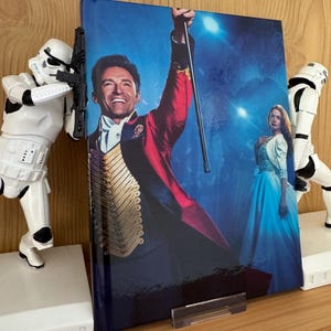 Puede incluir: Un libro de tapa dura con una imagen de un hombre con abrigo rojo y una mujer con vestido azul. El libro está sostenido por dos figuras blancas de Stormtroopers. El fondo es azul.