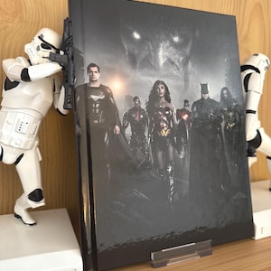 Puede incluir: Un libro de tapa dura con la Liga de la Justicia en la portada. La imagen muestra a Superman, Wonder Woman y Batman. El libro está sostenido por un soporte de acrílico transparente y figuras blancas de Stormtroopers a ambos lados.