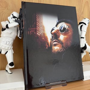 Puede incluir: Un libro de tapa dura con una imagen en primer plano de un hombre con gafas de sol, con un paisaje urbano de fondo. El libro está sostenido por dos figuras blancas de Stormtroopers, sobre una superficie de madera.