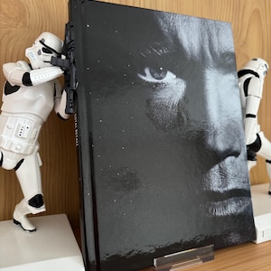 Puede incluir: Un libro negro con un primer plano de una cara en la portada, sostenido entre dos sujetalibros blancos de Stormtroopers. Los sujetalibros están sobre una superficie de madera. El libro se titula "TOTAL RECALL".