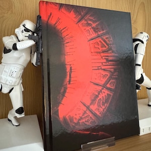 Puede incluir: Un libro de tapa dura con una cubierta brillante que presenta un gran diseño rojo sobre un fondo negro. Dos figuras blancas de Stormtroopers, que actúan como sujetalibros, flanquean el libro. El libro está sostenido por un soporte de plástico transparente.