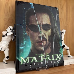 May include: A rectangular The Matrix Quadrilogy movie collection case featuring a portrait of a man with dark hair and sunglasses, set against a green digital code background. The case is flanked by two white plastic stormtrooper action figures.