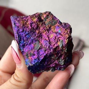 May include: A colourful, iridescent mineral specimen with a rough, textured surface. The stone displays a spectrum of colours, including deep purples, blues, and hints of gold and orange. The mineral is a natural, unpolished rock.