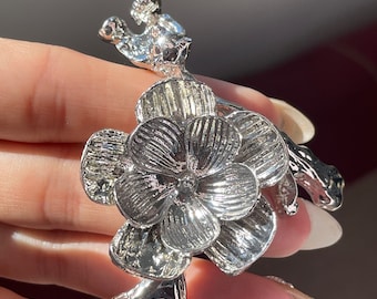 May include: Silver flower brooch with a detailed design. The flower has a centre with a raised, textured design and petals with a textured, ribbed design. The stem of the flower is twisted and has a detailed design.