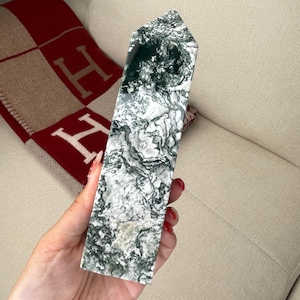 May include: A green and white marbled stone point, also known as a crystal, is held in a hand. The stone has a swirling pattern of green and white.
