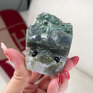 Puede incluir: Una piedra de ágata verde y blanca con dos agujeros negros perforados. La piedra es rugosa y tiene una forma natural y orgánica.