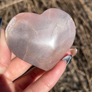 May include: A heart-shaped, polished stone in shades of pink and grey. The stone has natural veining and a smooth, reflective surface. The heart is held in a hand, with the background out of focus.