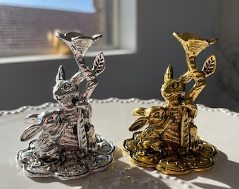 May include: Two decorative candle holders. One is silver, the other is gold. Each features a rabbit figurine holding a floral stem. The candle holders are on a white, scalloped plate.