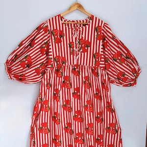 May include: A red and white striped dress with a strawberry print. The dress features long, puffed sleeves and a tie at the neckline. It is displayed on a wooden hanger.