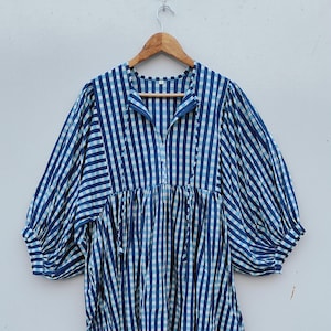 May include: A blue and white checkered dress with long, puffy sleeves and a gathered waist. The dress has a mandarin collar and a button-down front. The fabric appears to be lightweight, suitable for warmer weather. The dress is hanging on a wooden hanger.