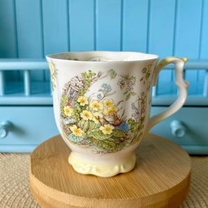 Puede incluir: Una taza de té de porcelana de color crema con una ilustración detallada de un pequeño animal rodeado de flores de prímula amarillas y follaje verde. La taza tiene un borde festoneado, una base de color amarillo pálido y un asa decorativa.