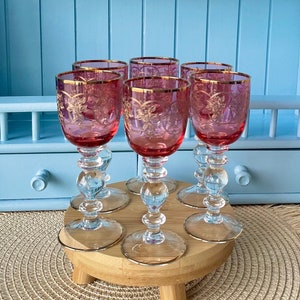 Può includere: Sei bicchieri da shot in vetro rosa e oro incisi su un supporto in legno. I bicchieri sono disposti in cerchio sul supporto. Il supporto è su un sottopentola in vimini.