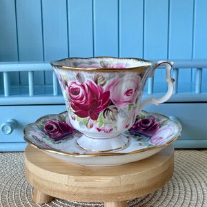 Può includere: Una tazza da tè e un piattino bianchi con un motivo floreale con rose rosa e rosse. La tazza da tè e il piattino sono su un supporto in legno con tre gambe.