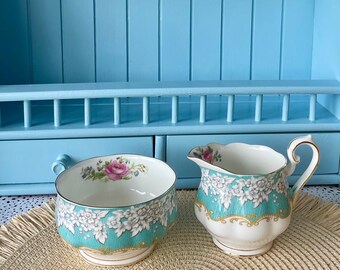 May include: A vintage porcelain sugar bowl and creamer set with floral and gold accents. The set is displayed on a light blue shelf with a woven placemat.