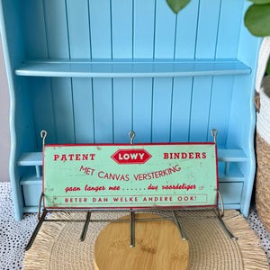 Puede incluir: Una estantería de pared de madera azul claro con dos estantes y un cajón pequeño. Un letrero vintage con el texto "LOWY BINDERS" cuelga debajo. Un objeto redondo de madera descansa sobre una estera tejida.