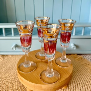 Könnte beinhalten: Satz von vier klaren Schnapsgläsern aus Glas mit goldenen und weißen Blumenmustern. Die Gläser sind auf einem Untersetzer aus Holz angeordnet.