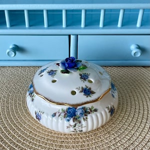 May include: White porcelain trinket box with a blue and white floral design and a blue rose on the lid. The box has a gold trim around the edge.