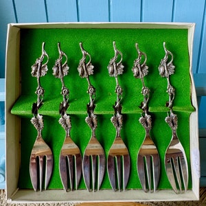 May include: Set of six silver-plated dessert forks with grapevine designs. The forks are in a white box with a green lining.
