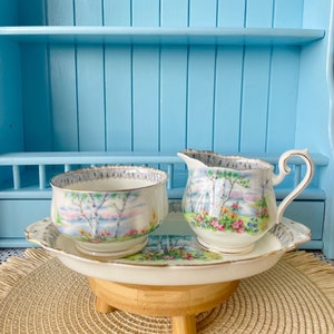 May include: A vintage porcelain sugar and creamer set with a floral and birch tree design. The set is on a white tray with a gold trim and is sitting on a wooden stand.