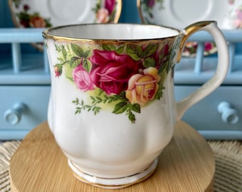 May include: A white teacup and saucer with a gold rim and a floral pattern featuring red, pink, and yellow roses. The cup has a handle and a scalloped edge.