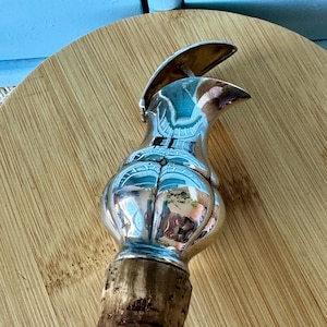 Può includere: Un tappo per bottiglia argentato con un design decorativo. Il tappo ha una base in sughero e una parte superiore dettagliata e riflettente. Il design include una forma curva, simile a un uccello. Il tappo è progettato per adattarsi a una bottiglia.