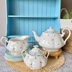 Può includere: Una credenza in legno azzurro espone un servizio da tè. Il set da tè comprende una teiera, una lattiera, una zuccheriera e piattini. I pezzi in porcellana bianca hanno un motivo floreale e finiture dorate. La teiera poggia su un piccolo supporto in legno.