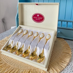 May include: A set of six gold-plated spoons with white porcelain handles, presented in a cream-coloured box. The interior lid features a red oval label with the text Royal Albert Val D'Or Gold Plated & Fine Porcelain. Each spoon is secured.