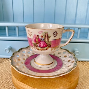 May include: A white teacup and saucer with a pink and gold design. The teacup has a pedestal base and features a painted scene of a couple in a garden. The saucer has a delicate openwork design.