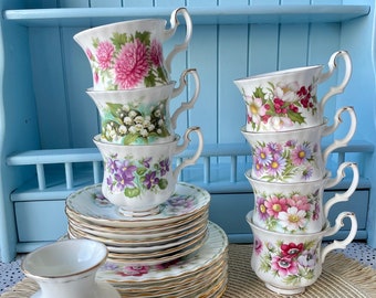 May include: A set of vintage teacups and saucers featuring floral designs. The cups are stacked in two piles, and the saucers are stacked in a pile. There is also a small vase with the word "February" on it.