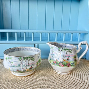 May include: A white porcelain sugar bowl and creamer set with a floral pattern featuring a birch tree landscape. The set has a gold trim and is decorated with a blue and gray border.