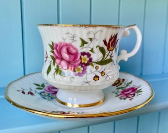 May include: A white teacup and saucer set with a floral design. The teacup features a large pink rose and other flowers, with gold trim on the rim and handle. The saucer has a matching floral pattern and gold trim. The set is on a light blue surface.