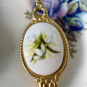 May include: A gold-tone spoon with a white porcelain oval-shaped handle featuring a floral design of white and green flowers.