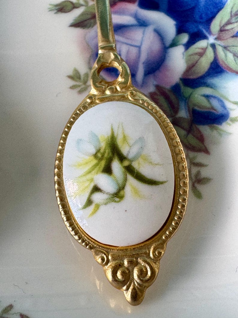 May include: A gold-tone spoon with a white porcelain oval-shaped handle featuring a floral design of white and green flowers.
