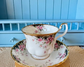 May include: A white teacup and saucer with a floral pattern in shades of pink, green, and gold. The cup and saucer have a scalloped edge and a gold trim.