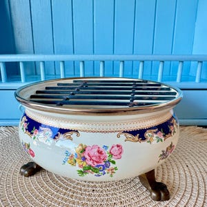 Puede incluir: Cuenco crema con decoración floral, borde azul oscuro y ribete dorado. El cuenco tiene una rejilla metálica y descansa sobre tres patas de color bronce. El diseño floral incluye rosas rosas y otras flores coloridas.