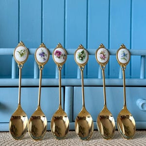 May include: Set of six gold teaspoons with floral designs on the handles. Each spoon features a different floral pattern in shades of pink, purple, and white.