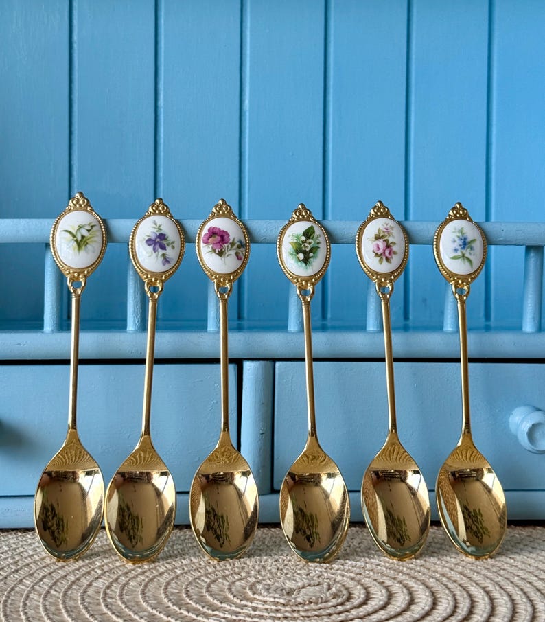 May include: Set of six gold teaspoons with floral designs on the handles. Each spoon features a different floral pattern in shades of pink, purple, and white.