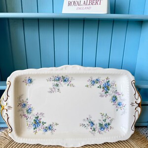 May include: A rectangular white porcelain serving tray featuring a delicate floral pattern of blue roses and green leaves. The tray has scalloped edges with gold trim on the handles. A Royal Albert England sign is positioned above the tray.