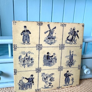 May include: A square, off-white tile panel featuring nine blue and white Dutch-style illustrations. The images depict scenes with people, windmills, and floral motifs. The tile has a slightly aged appearance, with visible cracks and imperfections.