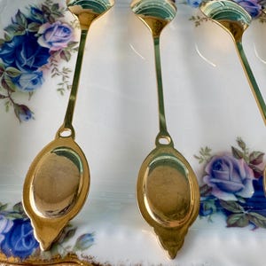 May include: Four gold-coloured teaspoons with ornate handles are arranged on a white porcelain plate with a floral pattern. The spoons are polished and reflect light.