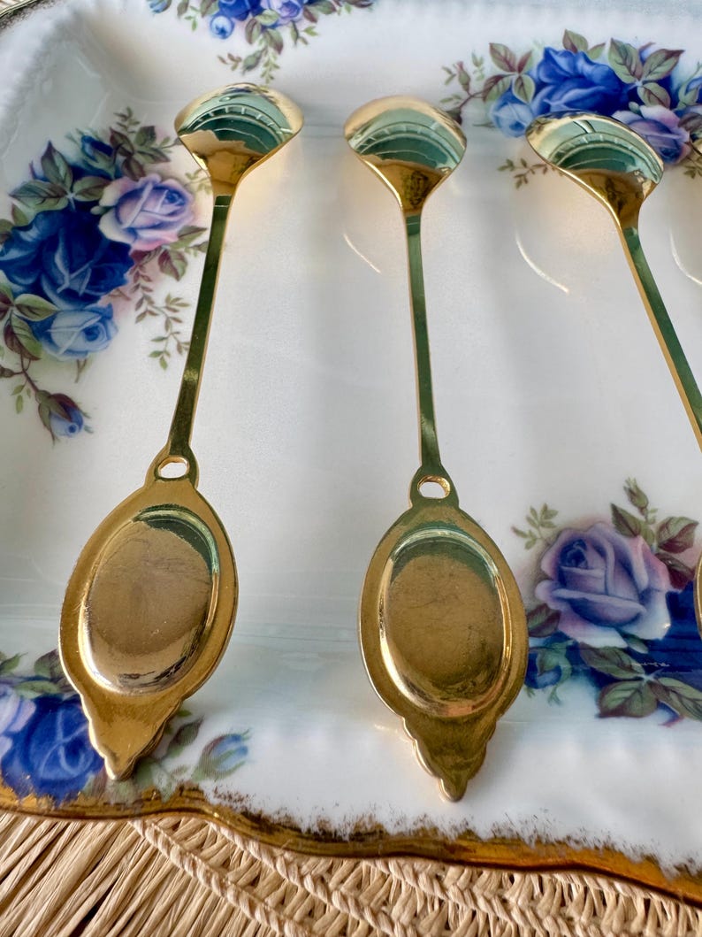 May include: Four gold-coloured teaspoons with ornate handles are arranged on a white porcelain plate with a floral pattern. The spoons are polished and reflect light.