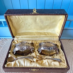 May include: An antique silver salt cellar set with two ornate bowls and spoons, nestled in a brown, textured case with a gold satin lining. The silver pieces have floral detailing. The case is closed with a gold clasp.