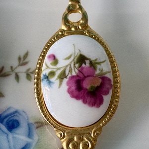 May include: A gold-tone spoon with a white porcelain oval-shaped handle featuring a floral design with pink and purple flowers and green leaves.