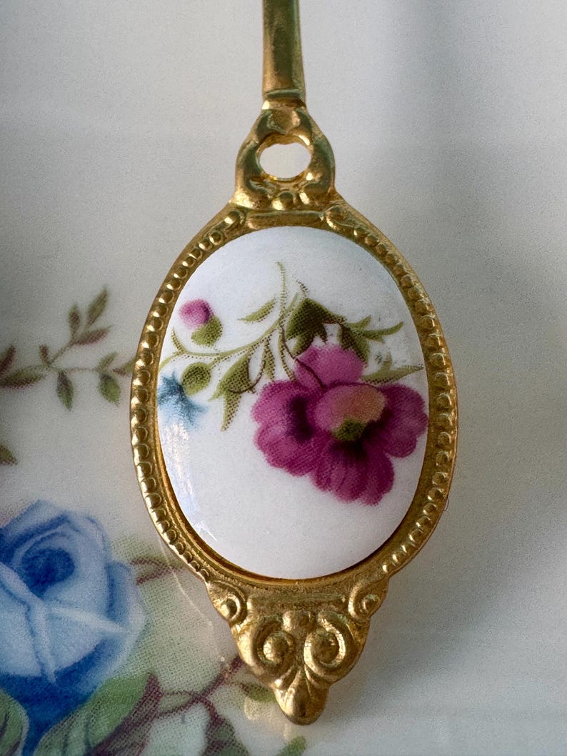 May include: A gold-tone spoon with a white porcelain oval-shaped handle featuring a floral design with pink and purple flowers and green leaves.
